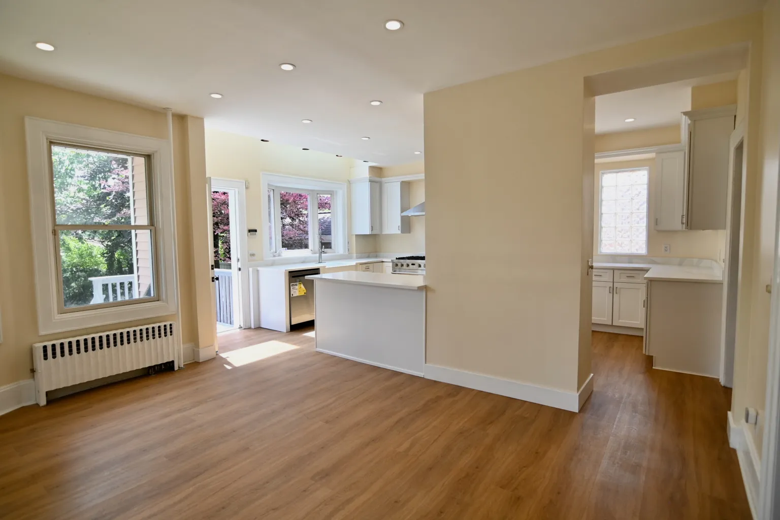 Looking from the dining room across the open floor into the kitchen, a tall window and cast-iron radiator at left and an opening through to the pantry at right