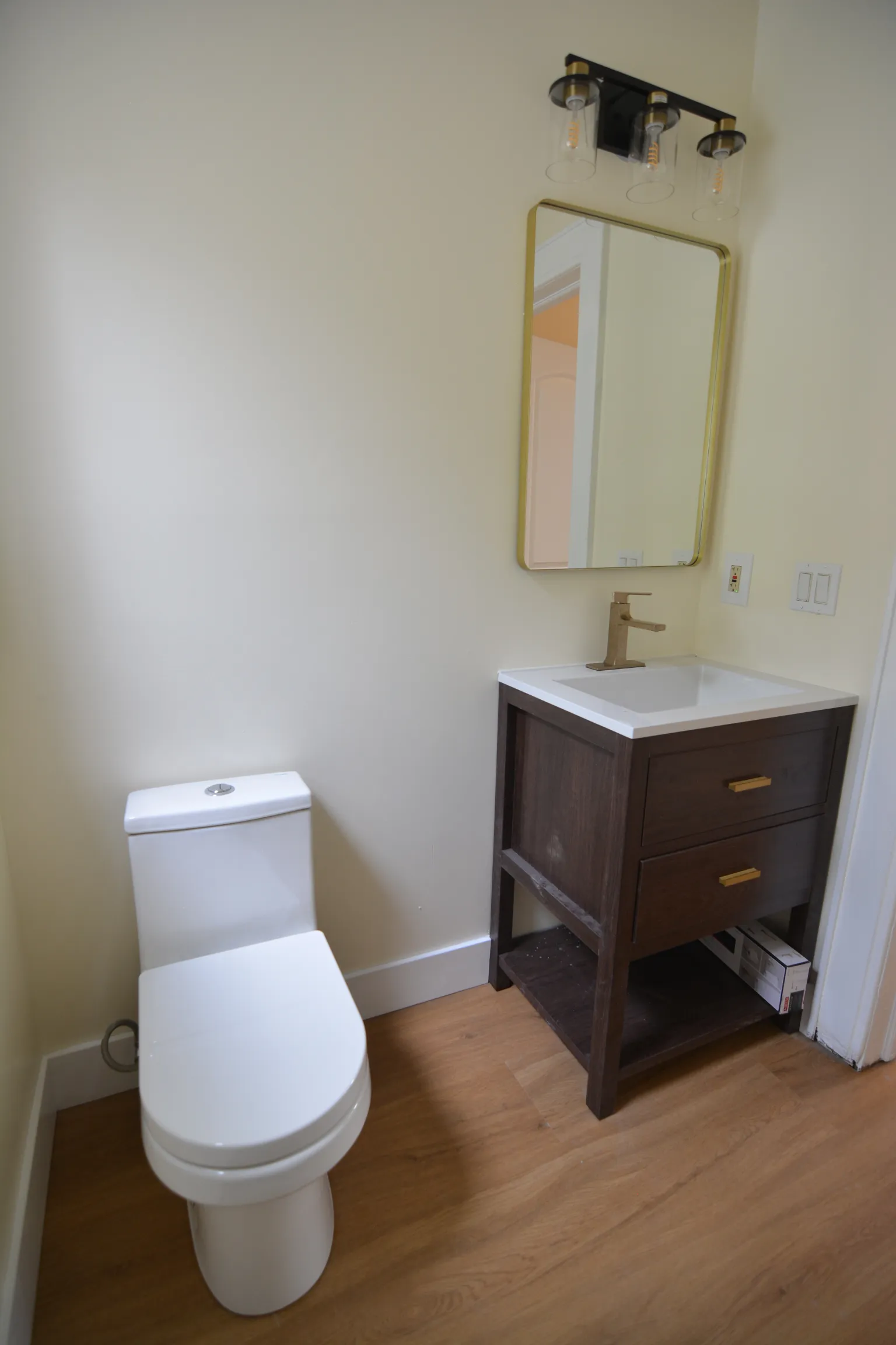 Renovated main-floor half bath with a dark-wood open-frame vanity, brass-framed mirror, Edison-bulb sconces, and a modern toilet