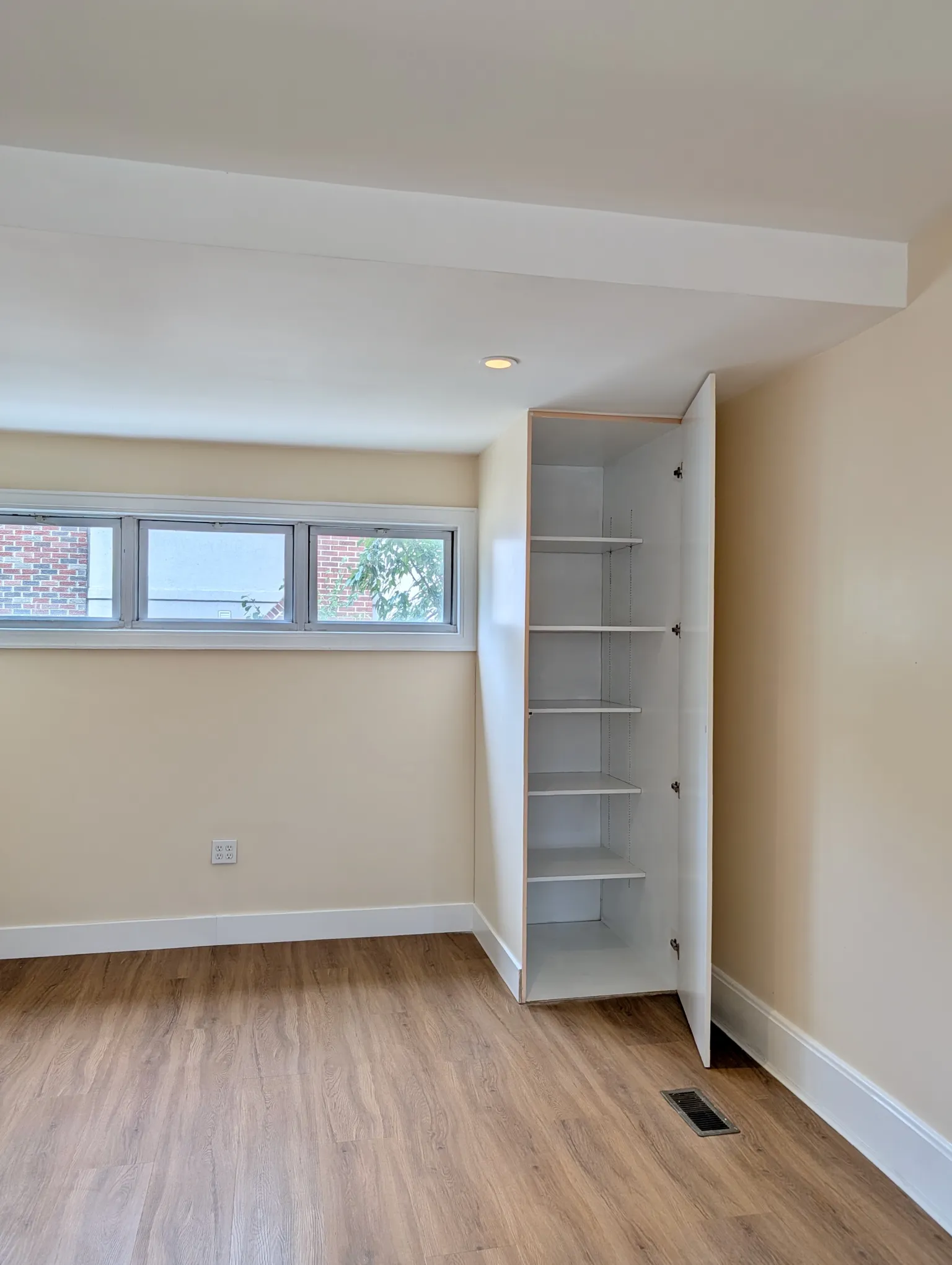 A tall built-in closet at the far end of the living room, door open to reveal full-height shelving, set beneath the high windows