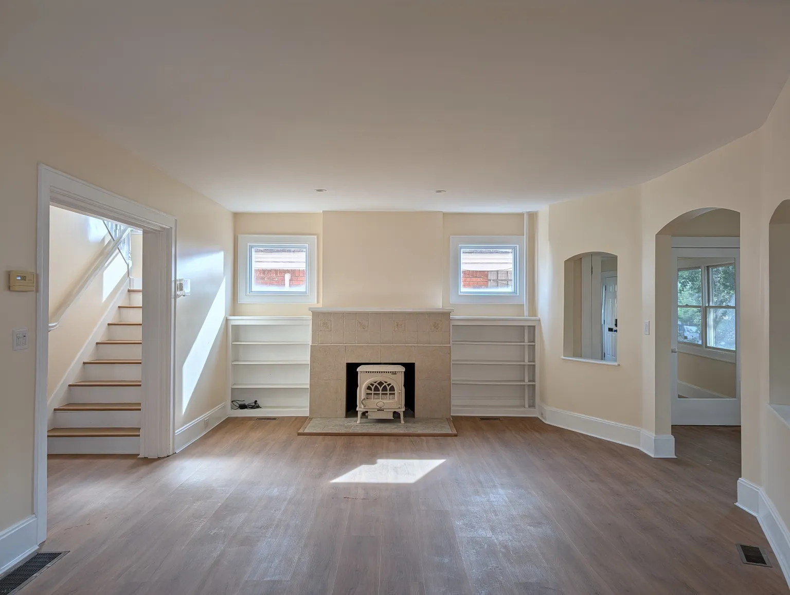 The main-floor living room with the enameled stove set into a tiled hearth between symmetric built-in shelves, twin windows above, the staircase on one side, and an arched opening through to the entry on the other