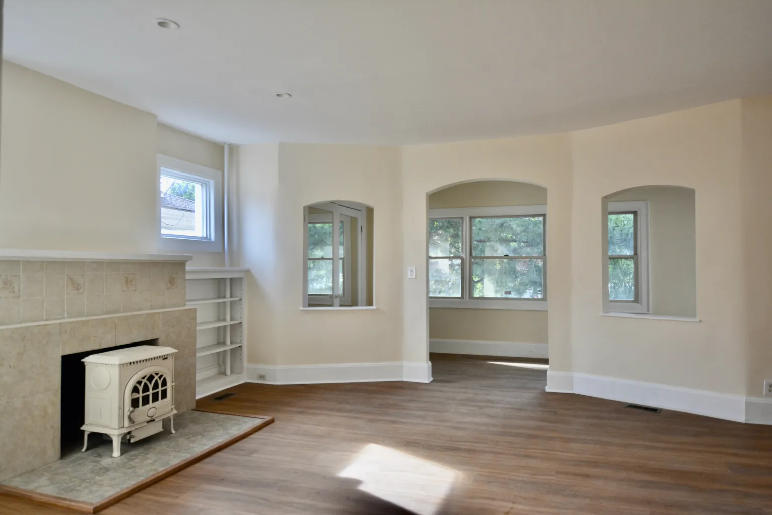 The living room with the cream enameled wood stove set into a tiled hearth beside built-in shelves, and a curved bay wall with three arched windows looking out to the trees