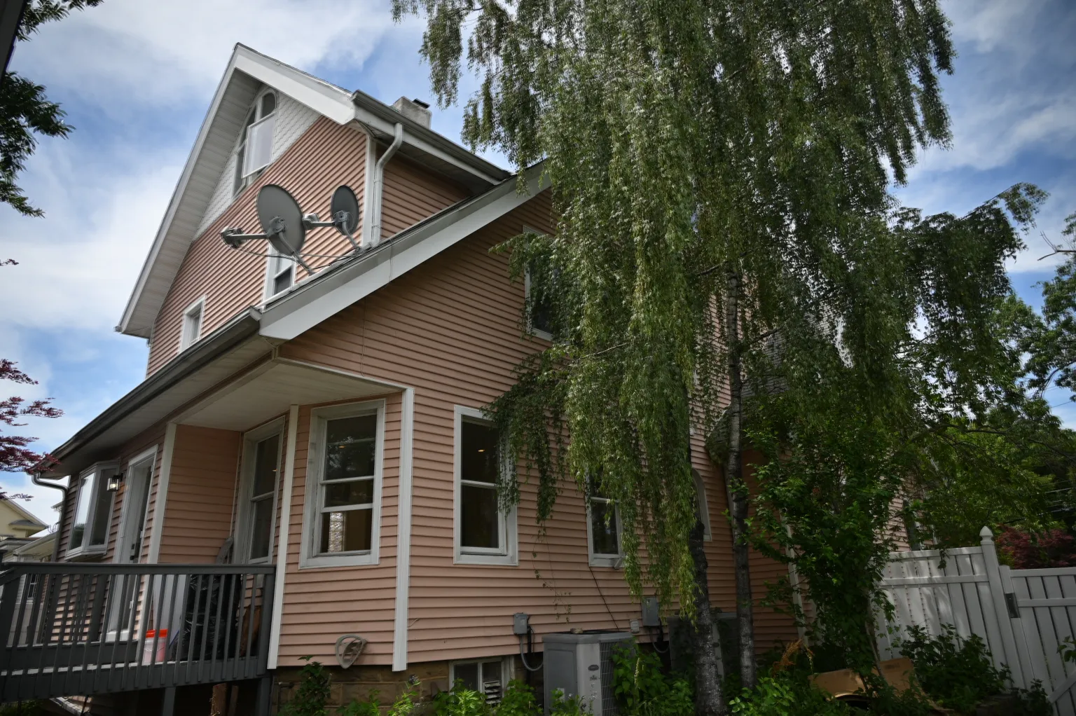 The peach-sided side of the house under its cross-gable roof, a large tree draping over the side yard, with the deck at left and a white vinyl fence along the property line