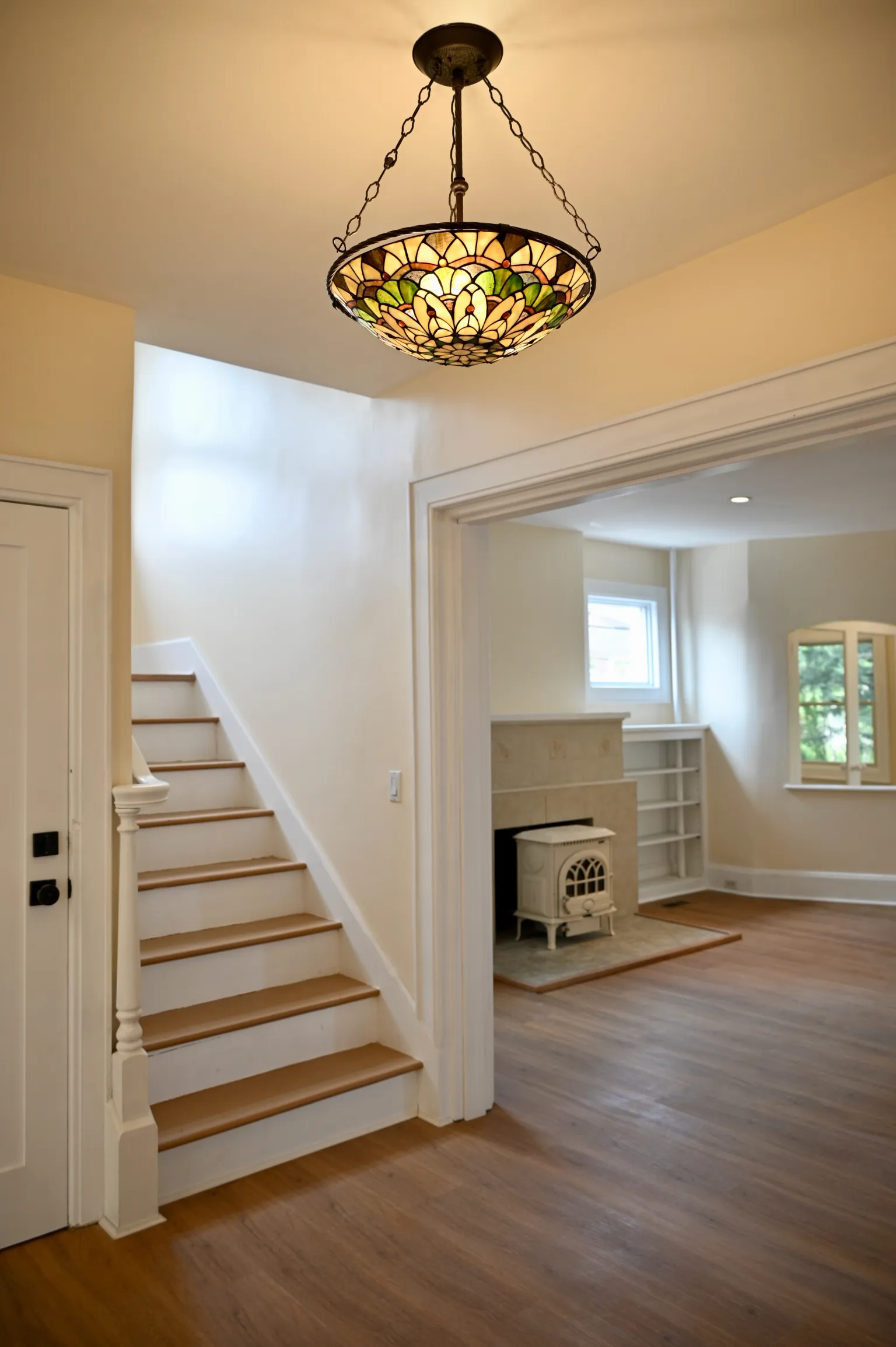 The main-floor staircase rising to the second floor, a stained-glass pendant light overhead, with an archway through to the living room and its enameled stove beyond
